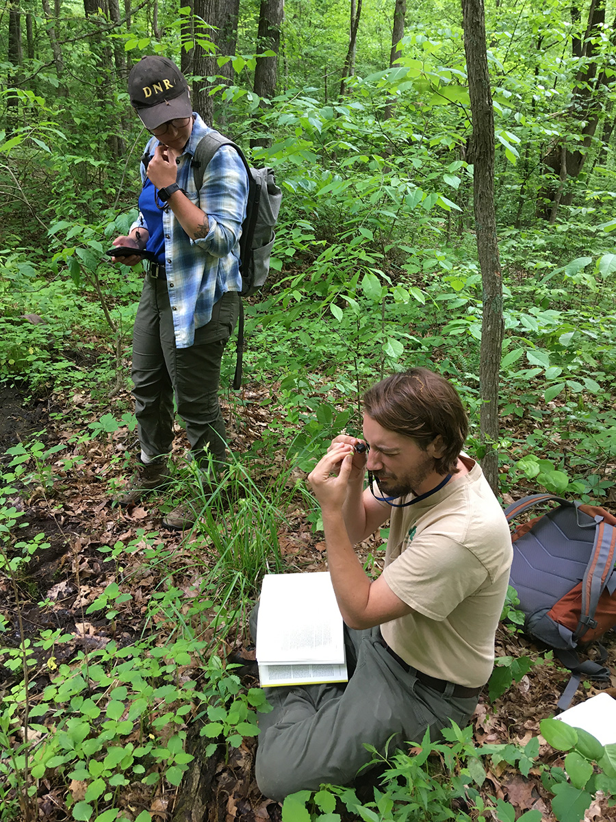 Two DNR ecologists conduct a bioinventory survey on a wooded property in Southern Indiana. In the foreground a man sits on the forest floor looking closely at a specimen through a magnifying loupe while in the background another biologist looks towards him.